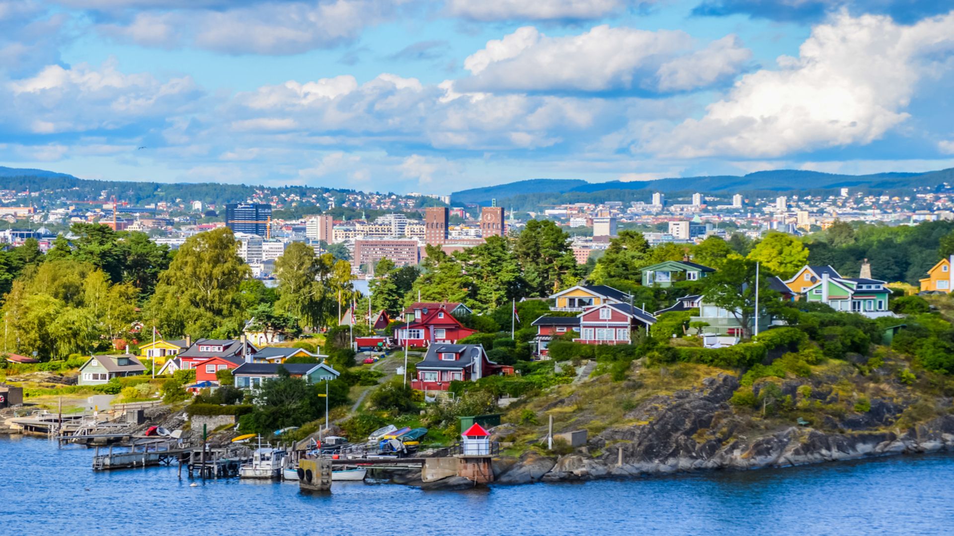 Colourful wooden cottages on a rocky shore in the Oslofjord, with Oslo's city hall on the skyline behind
